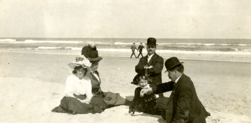 People in Edwardian clothing sitting on a beach.